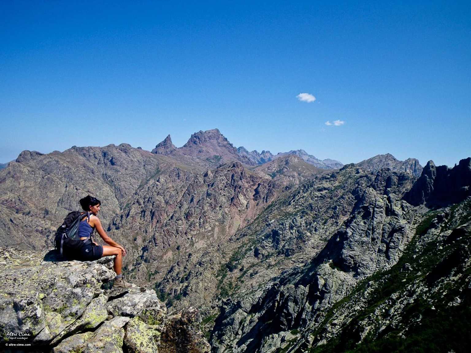 Randonneur se reposant sur un rebord rocheux surplombant le terrain montagneux accidenté du sentier GR20 en Corse sous un ciel bleu clair